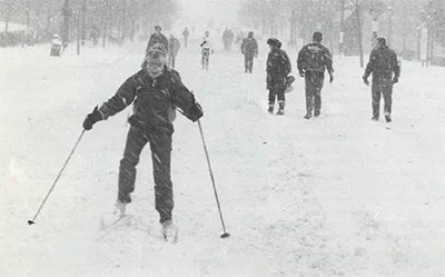 Black and white photo of woman cross-country skiing down the street after the 1991 Halloween blizzard. Eight other people are seen walking in the opposite direction.